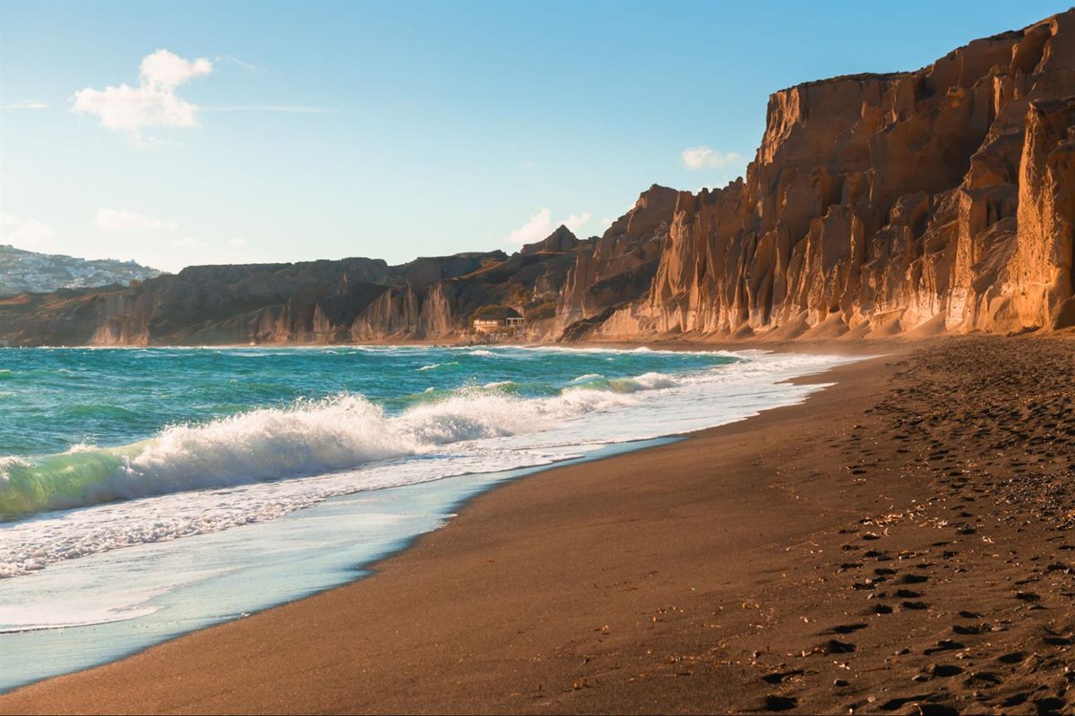 Waves breaking on Vlychada's black sand beach beneath Santorini's cliffs at golden hour